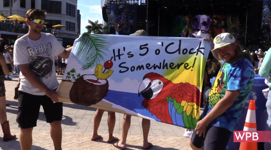 two guys hold a banner up outside that says " it's 5 O'clock Somewhere" and there's a stage behind them