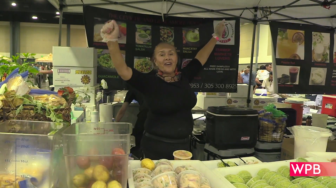 a woman holds her hands in the air mid dance, smiling at a booth full of mangoes during the Florida Mango Festival 