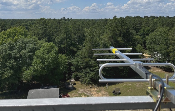 Silver antenna pointing out, from high on tower, over green tree tops and toward rim of white-blue sky 