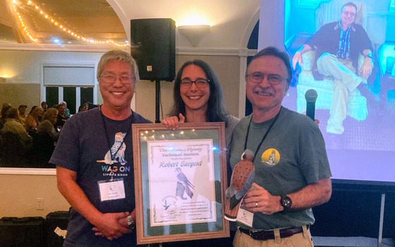 Three people standing and holding a large plaque with crowd in the part of the background