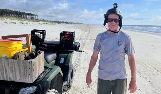 Man in gray T-shirt with black head gear on white sand beach with ATV