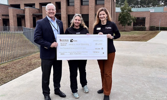 Three people holding white, poster-sized check on gray sidewalk in front of orange-brick school buildings