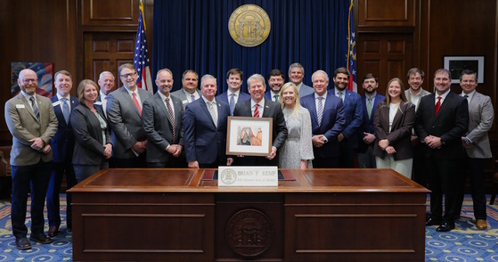 Group including Gov. Kemp and First Lady behind dark wood desk against dark blue and brown background