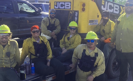 7 DNR staff in yellow fire shirts smudged with black soot rest on a trailer bed