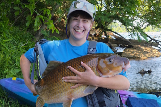 Woman in blue shirt and DNR cap holds large golden-brown fish with river bank and water in background