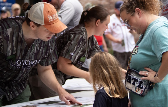 DNR staff in camo jerseys talk with a little girl and adult woman at a DNR table