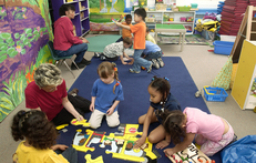 Two small groups of preschool teachers and preschool children playing on classroom carpet. 