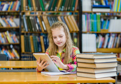 Girl sitting at library table surrounded by books and holding an electronic tablet.