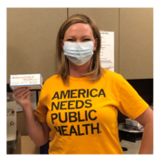 Amy posing in her America Needs Public Health shirt with a box of COVID-19 vaccine