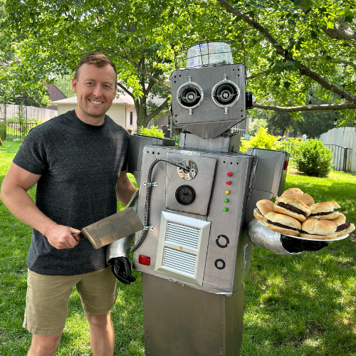 A man stands beside Grillbert 9000, the BBQ robot, who holds a sandwich, ready to share grilling tips with humor.