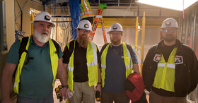 Four workers in safety vests and hard hats stand in a construction area with exposed wiring, ladders, and equipment.