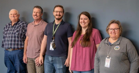 Five people stand side by side against a plain wall, wearing casual and workplace attire with ID badges visible