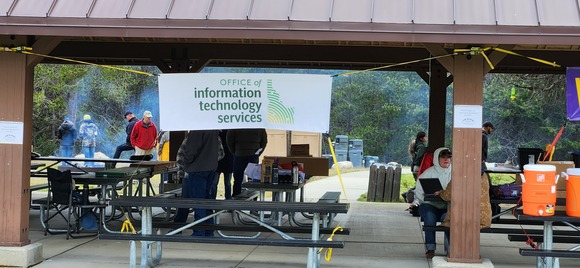 Outdoor pavilion with an Information Technology Services banner, picnic tables, people gathered, equipment and coolers set up