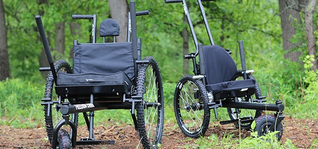 photograph of two all-terrain wheelchairs on an unpaved path in a forest setting