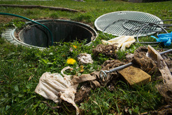 Open ground utility hole in a grassy area with its cover set aside, surrounded by debris 