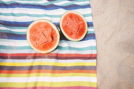 beach towel on the sand with a watermelon cut in half sitting on the towel