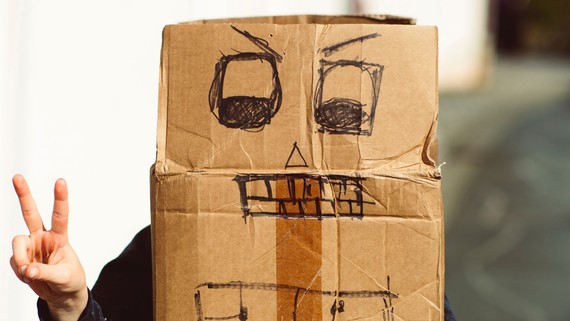 A kid wearing a cardboard box with a crazy face drawn on it holds up a peace sign.