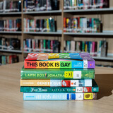 Stack of 5 banned books on a library table. The top book is "This Book is Gay" and it has a rainbow cover