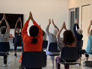 Roomful of people sitting in chairs with arms raised, sitting with backs to the camera, and one person in front leading class