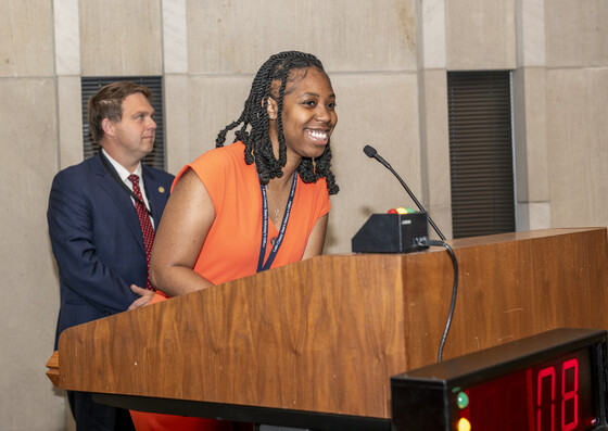 Woman in orange suit speaking behind a podium 