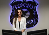A woman, Samantha McCabe of the Peoria Police Department, stands in front of a Peoria Police badge neon sign.
