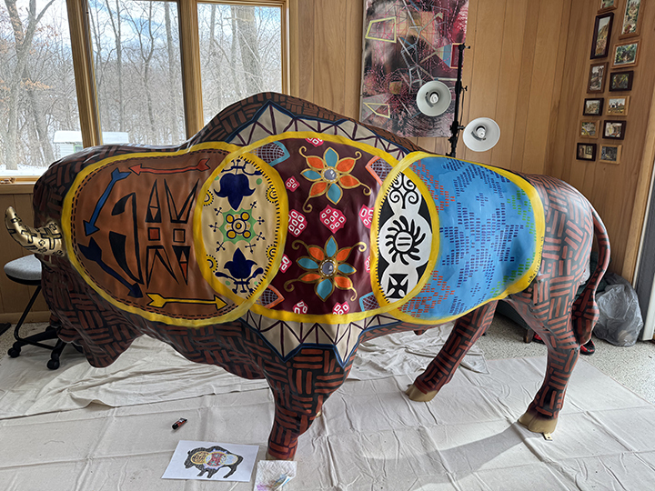The finished product of the painted bison sculpture stands in artist's basement, with a drawing of the design on the ground.