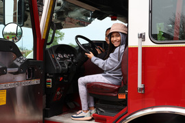 A young girl sits with her hands on the wheel of a fire apparatus while another child peers around her.