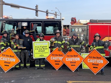 Firefighters in gear line up in front of a red fire truck with signs about the Fill the Boot charity.