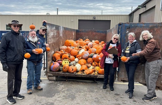 Group of men and women standing next to a dumpster filled with peoples' discarded pumpkins that are ready for composting.