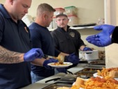 Three firefighters spoon pasta onto plates in the kitchen.