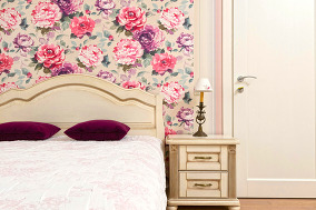 A bed and nightstand next to a closed door; floral wallpaper and pink and purple accents.