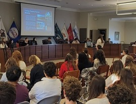students are seated in council chambers with a screen showing a projected powerpoint presentation.