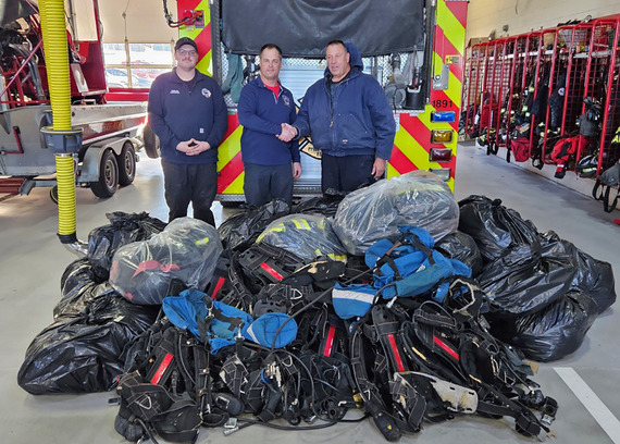 Three men shaking hands in the fire station with a pile of donated gear on the ground.
