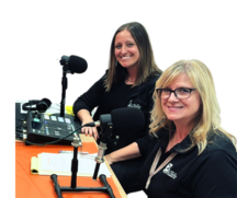 Two women sitting at a desk with a soundboard and microphones