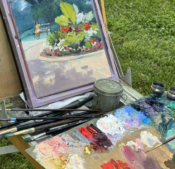 A painting on an easel with a person dipping a brush into a color on a palette.