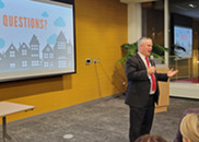 The mayor speaks to an audience with a presentation on a big white screen behind him.