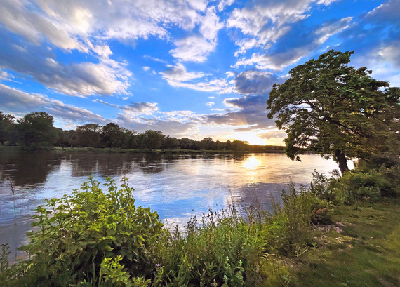 The sun sets in the west while the Fox River flows by, with greenery and trees surrounding its banks.