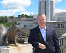 Mayor Clint Hull stands along the riverfront, next to a pair of fox statues with the municipal building in back.