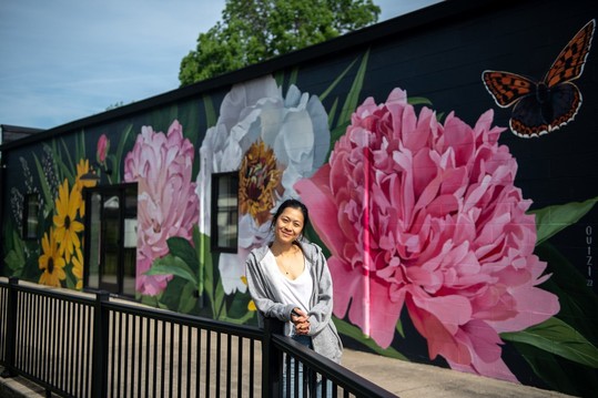 Louise Jones standing in front of her mural of local flowers including peonies and black-eyed susans painted largely on a black brick wall.