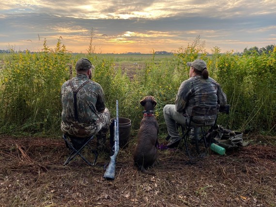 Two hunters dressed in camouflage sit with a dog between them.