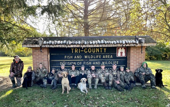 Thirteen youth hunters dressed in camouflage and four dogs sit in front of a Tri-County FWA sign.