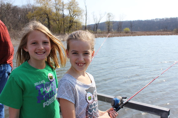 Two children standing by a body of water as one of them holds a fishing pole.
