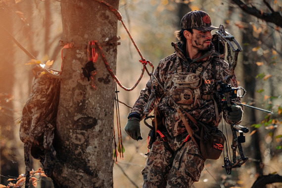 A bow hunter standing in the woods while hunting from a saddle.