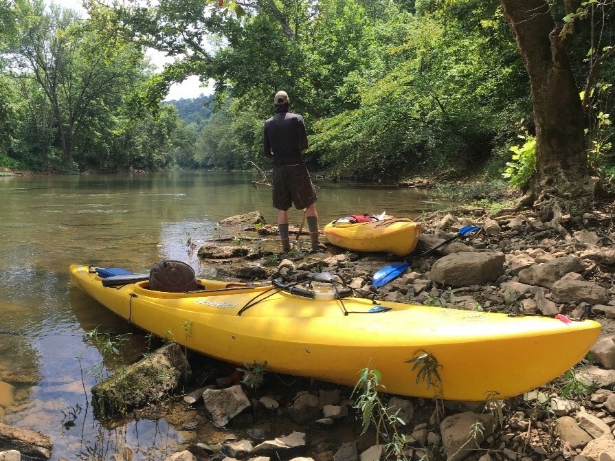 A man shoreline fishing beside a kayak in the woods.