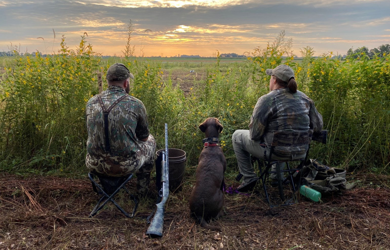 two hunters dressed in camouflage sit with a dog