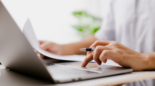 Hand on the trackpad of a laptop, also holding pen, other hand is holding a document