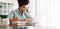 Woman sitting at a table, looking at a laptop and writing in a notebook