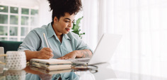 Woman sitting at a table, looking at a laptop and writing in a notebook