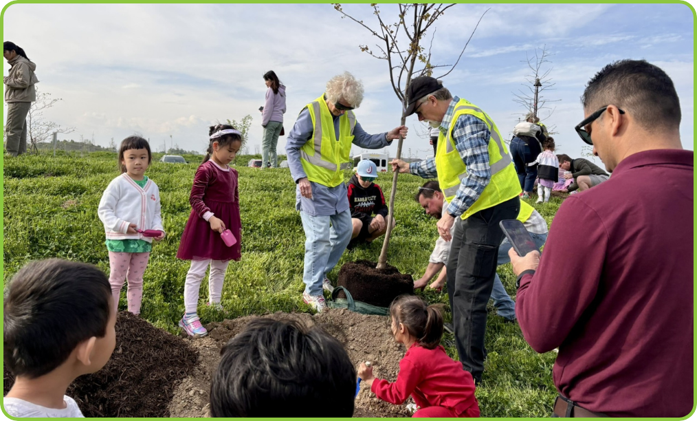 Two tree frined volunteers in safety vests helping plant a sapling tree at a hole surrounded by children