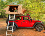 A Young Boy Pops His Head Out of a Camper On Top of a Red Jeep in the Middle of the Woods